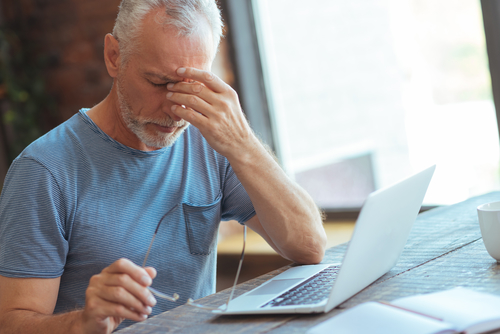 A man rubs his forehead in frustration while reviewing his investment accounts on his laptop.