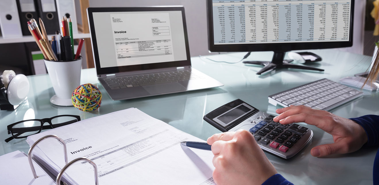 A business owner working at their desk with computer, laptop and calculator reviewing cash flow. 